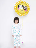 Child wearing glasses and a white outfit with cloud patterns, holding a sun-shaped balloon with sunglasses and pink cheeks.